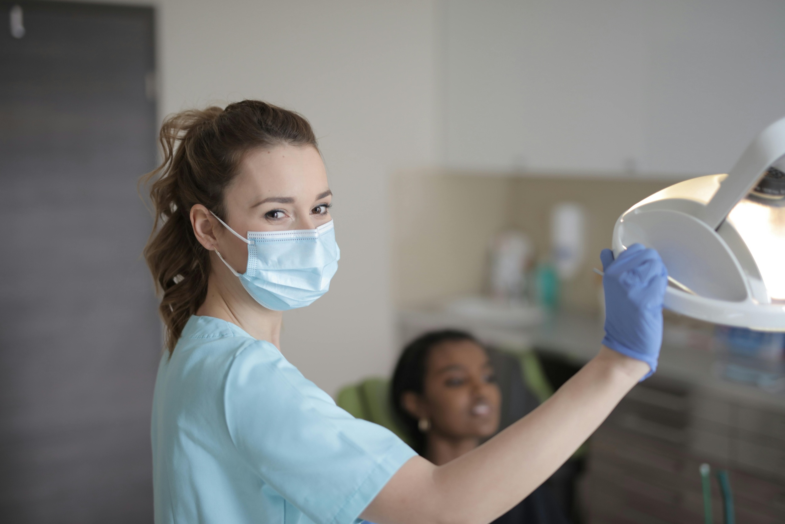 Behandlerin arbeitet mit Maske im Behandlungsraum, hat eine Hand an der Lampe.